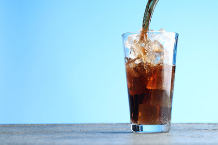 Pouring refreshing cola into glass at grey textured table against light blue gradient background, closeup. Space for textの写真素材