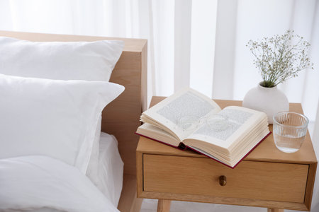 Bedside table with book, flowers in vase, glass of water and glasses near bed indoorsの写真素材