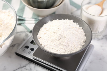 Digital kitchen scale with bowl of flour and other products on white marble table, closeupの写真素材