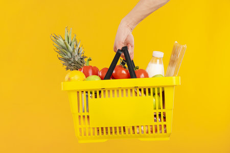 Man holding shopping basket with different products on yellow background, closeupの写真素材