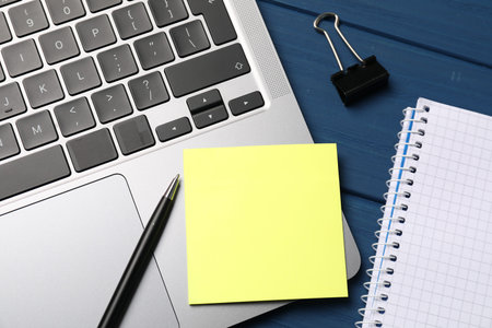 Laptop with sticky note, pen, paper clip and notebook on blue wooden table, closeup