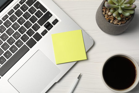 Sticky note, pen, coffee, floral decor and laptop on white wooden table, flat lay