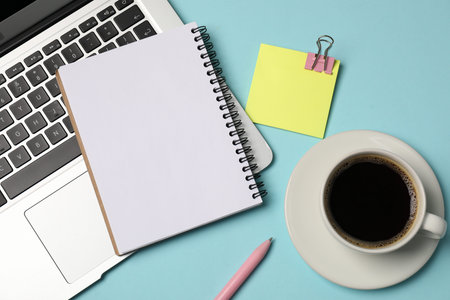 Notebook, sticky note, pen, coffee and laptop on light blue background, flat lay