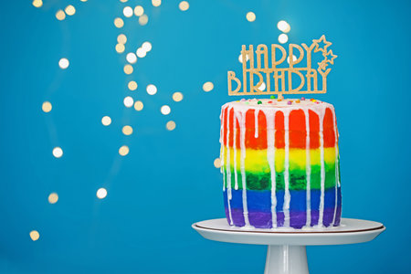 Delicious rainbow birthday cake with topper on stand against light blue background with blurred lights, closeup. Space for textの写真素材
