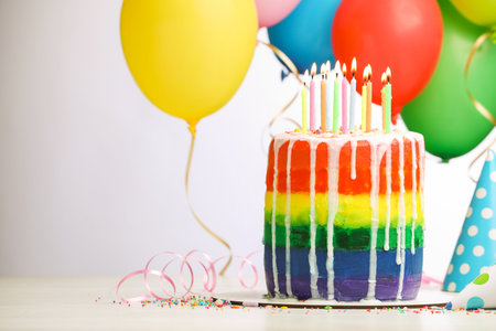 Delicious rainbow birthday cake with burning candles, streamers, sprinkles, party hats and balloons on white table against light background, closeup. Space for textの写真素材