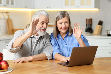 Cute elderly couple having video call by laptop at table in kitchen