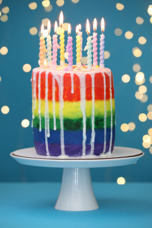 Delicious rainbow birthday cake with candles on light blue table against blurred lights, closeup