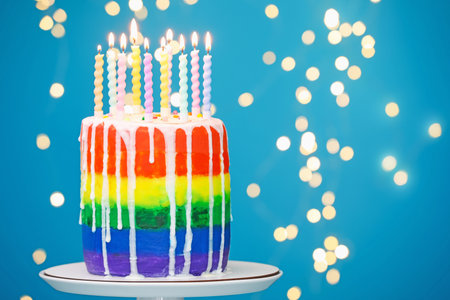 Delicious rainbow birthday cake with burning candles on light blue background with blurred lights, closeup. Space for textの写真素材