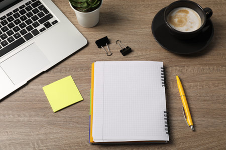 Notebook, pen, sticky note, binder clips, coffee, floral decor and laptop on wooden table, above...