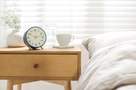 Bedside table with alarm clock, flowers in vase, cup and glass of water near bed indoors, closeup