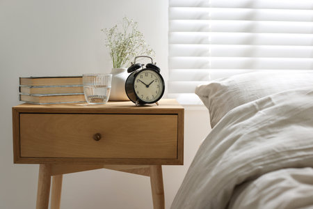 Bedside table with books, flowers in vase, alarm clock and glass of water near bed indoors
