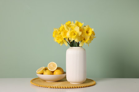 Beautiful daffodils in vase and lemons on white table near light green wallの写真素材