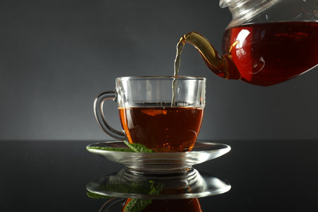 Pouring freshly brewed tea from teapot into cup at dark mirror table, closeupの写真素材