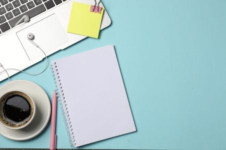 Notebook, pen, sticky note, earphones, coffee, binder clip and laptop on light blue background, flat...