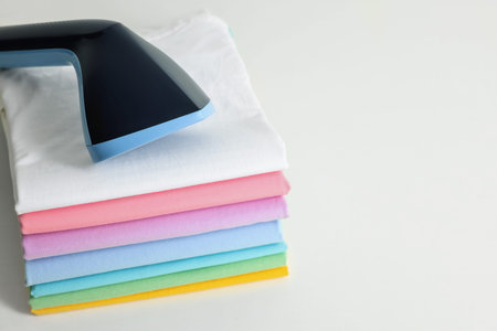 Upright steamer and stack of folded clothes on white table, closeup. Space for text