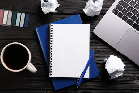 Open blank notebook, other stationery, coffee, crumpled paper and laptop on black wooden table, flat...