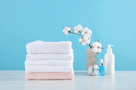 Stack of clean towels, cotton flowers and cosmetic products on white marble table against light blue backgroundの写真素材