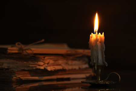 Burning candle in candlestick and old books on mirror surface in darkness, closeupの写真素材