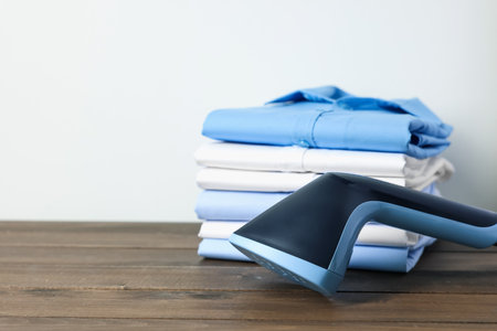 Upright steamer and stack of folded shirts on wooden table near white wall indoors, closeup. Space for textの写真素材