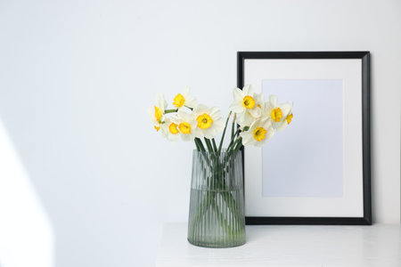 Beautiful daffodils in vase and picture frame on white wooden table indoors, space for textの写真素材