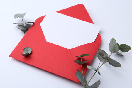 Blank greeting card in red paper envelope and eucalyptus leaves on white background, closeup