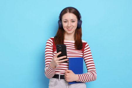 Teenage girl in headphones with smartphone and notebook on light blue background