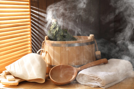 Sauna equipment. Bucket, ladle, felt wool hat, whisk and towels on bamboo bench in steamの写真素材