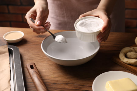 Making pretzels. Woman adding baking soda into bowl of water at wooden table, closeupの写真素材
