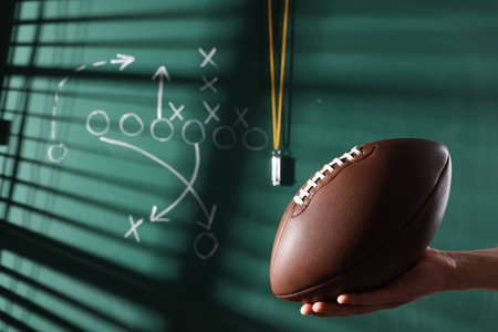 Man holding American football ball near chalkboard with scheme of game, closeup