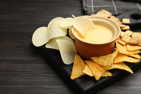 Tasty cheese dipping sauce in bowl and different snacks on dark wooden table, closeup. Space for textの写真素材