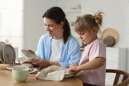 Little girl helping her mom wiping plates at table in kitchenの写真素材
