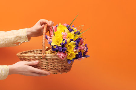 Woman holding wicker basket with beautiful flowers on orange background, closeup. Space for textの写真素材