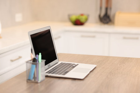Laptop with blank screen and stationery on wooden table in kitchen, selective focus. Mockup for designの写真素材