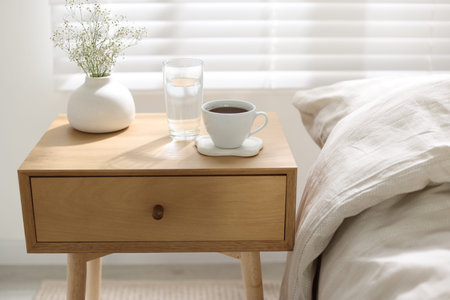 Bedside table with glass of water, flowers in vase and coffee near bed indoors, closeup