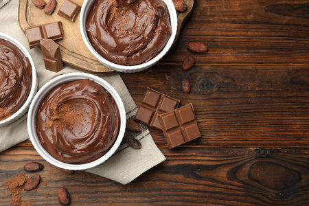 Tasty pudding in bowls, pieces of chocolate and cocoa beans on wooden table, flat lay. Space for...