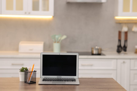 Laptop with blank screen, houseplant and stationery on wooden table in kitchen. Mockup for designの写真素材