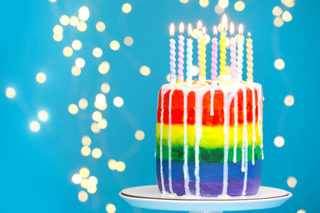 Delicious rainbow birthday cake with burning candles on light blue background with blurred lights, closeup. Space for textの写真素材