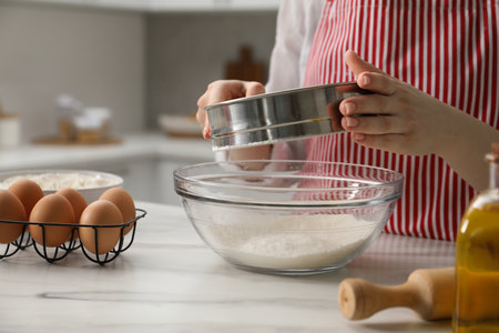 Making khinkali. Woman sieving flour into bowl at table in kitchen, closeupの写真素材