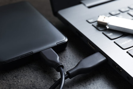 USB flash drive, hard disk drive with cable and laptop on dark grey table, closeup
