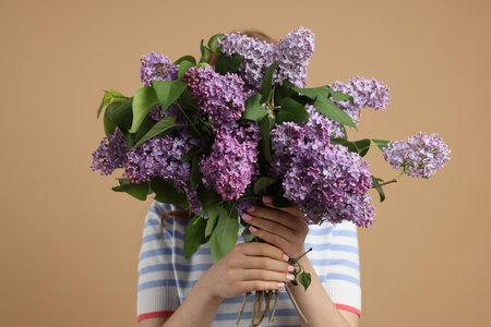 Teenage girl hiding behind bouquet of lilac flowers on beige backgroundの写真素材
