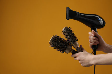 Woman with hair dryer and round brushes on orange background, closeup. Space for textの写真素材