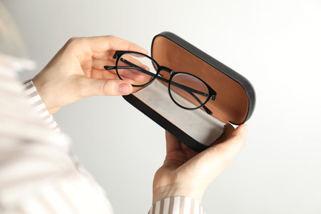 Woman holding case with stylish glasses and cleaning cloth on light background, closeup