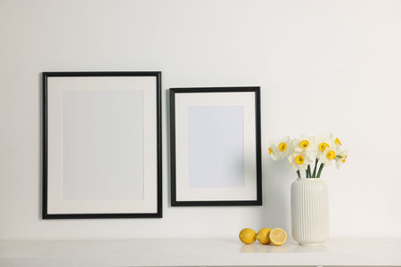 Beautiful daffodils in vase on white table and picture frames indoors