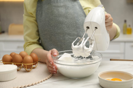 Woman whisking cream with hand mixer at white marble table indoors, closeup