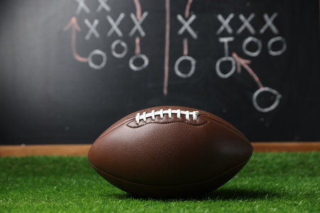 American football ball on green grass against chalkboard with game schemeの写真素材