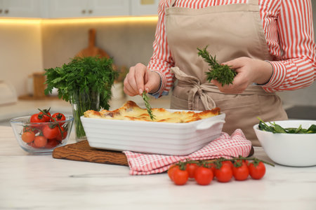 Woman putting rosemary onto lasagna at white marble table in kitchen, closeupのeditorial素材