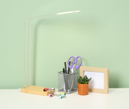 Stylish workplace with lamp, stationery and plant on white desk near light green wallの写真素材