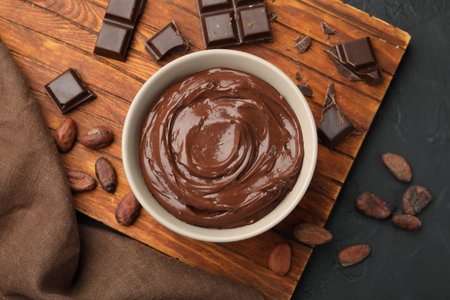 Tasty pudding in bowl, pieces of chocolate and cocoa beans on black table, flat layの写真素材
