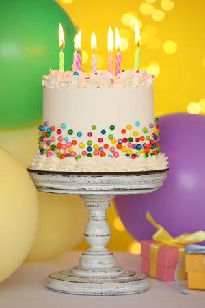 Tasty cake with colorful sprinkles, burning candles and other festive decor on table against yellow background, bokeh effectの写真素材