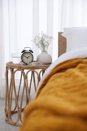 Bedside table with alarm clock, flowers in vase, glass of water and glasses near bed indoors